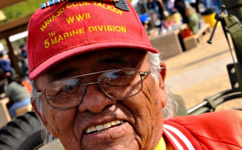 A Veteran Navjao Code Talker smiles at the camera. He is wearing a red hat that says Navajo Code Talker WWII 5th Marine Division.
