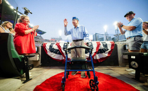 Veteran Waitzman stands in front of banners of flags. He is waving. People around him are clapping. The stadium lights are on behind him.