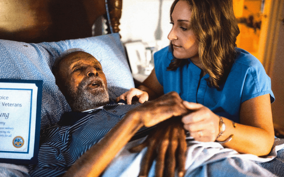 A health aide holds the hands of a Veteran lying in bed.