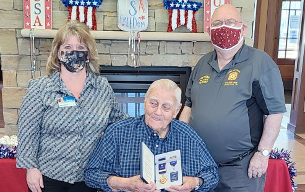 Mr. Allen, sitting in a chair, holds his Korean War Peace Medal. Two Caris Healthcare staff members are behind him.