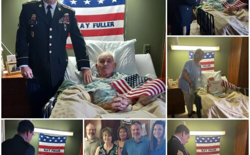 Ray Fuller is in his bed as a service member and family stand around him smiling. A flag is on the wall that says Ray Fuller and has stars and stripes around it.