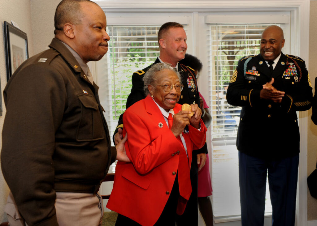 An older Black woman vVteran smiles as she receives a medal. Service members stand around her clapping and smiling.